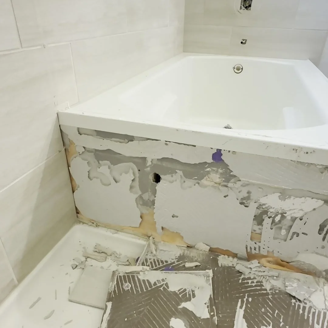 A bathtub during demolition with the tile stripped from its lower face and removed tiles lying on the floor