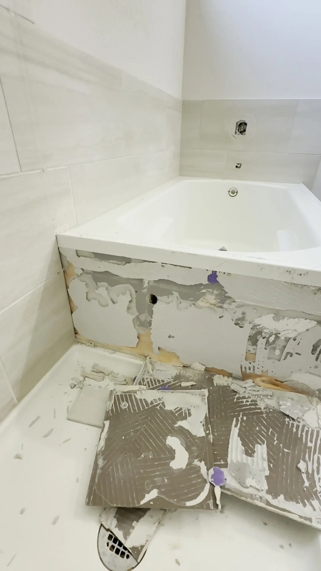 Removed bathroom tiles lying face down on the floor beside a bathtub, their backs showing combed thinset ridges with bare patches, and stripped drywall behind them