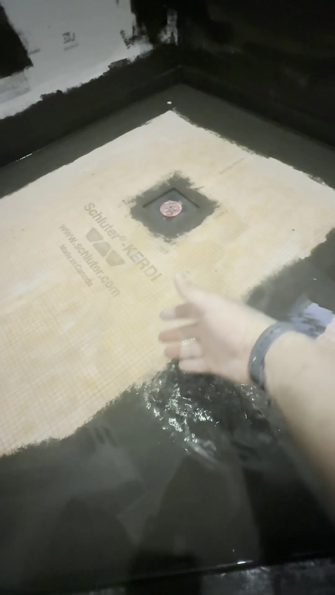 A shower pan filled with water during a flood test, the plugged drain visible beneath the surface and dark waterproofing on the walls, with a hand dipped into the water