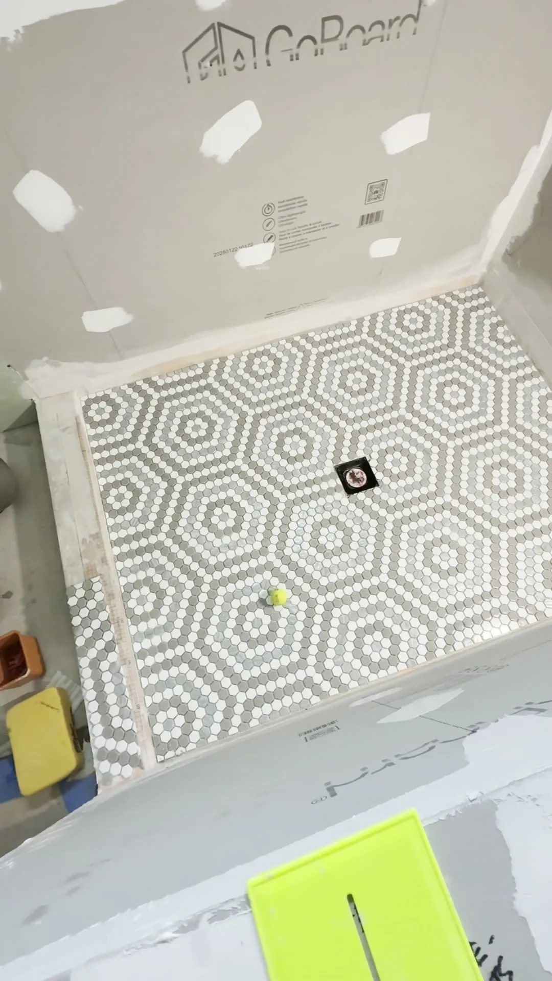 Looking down into a shower pan with a completed floor of small grey and white hexagon mosaic laid in a concentric-hexagon pattern, the square drain cut out at the centre and unfinished waterproof backer board on the walls