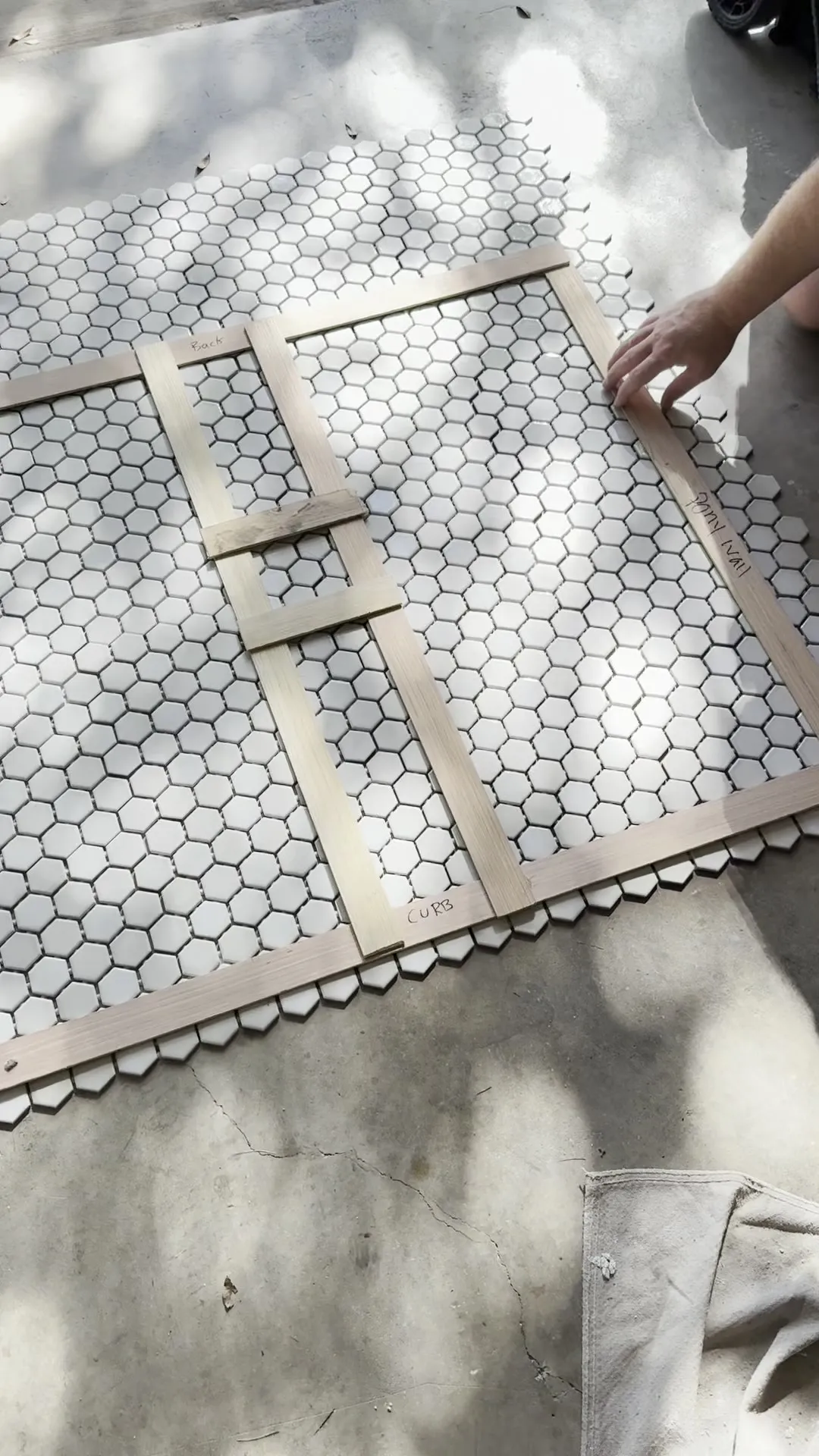 A frame of thin wooden strips, hand-labelled with the positions of the shower's back wall, pony wall and curb, laid over sheets of white hexagon mosaic tile on concrete outdoors, with a hand adjusting one strip