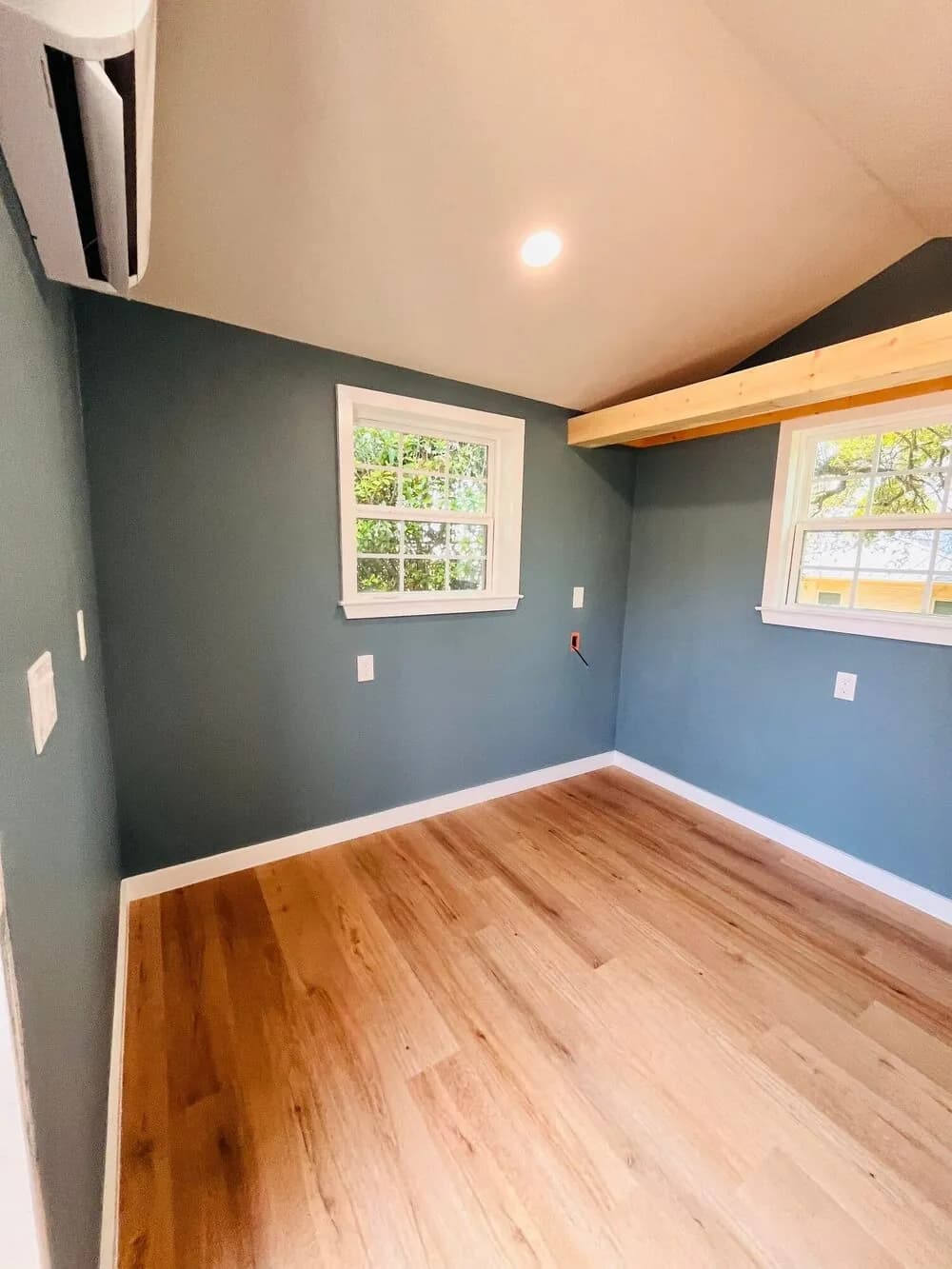Interior of a finished studio conversion with a wall-mounted mini-split above plank flooring, beside a window and an exposed beam