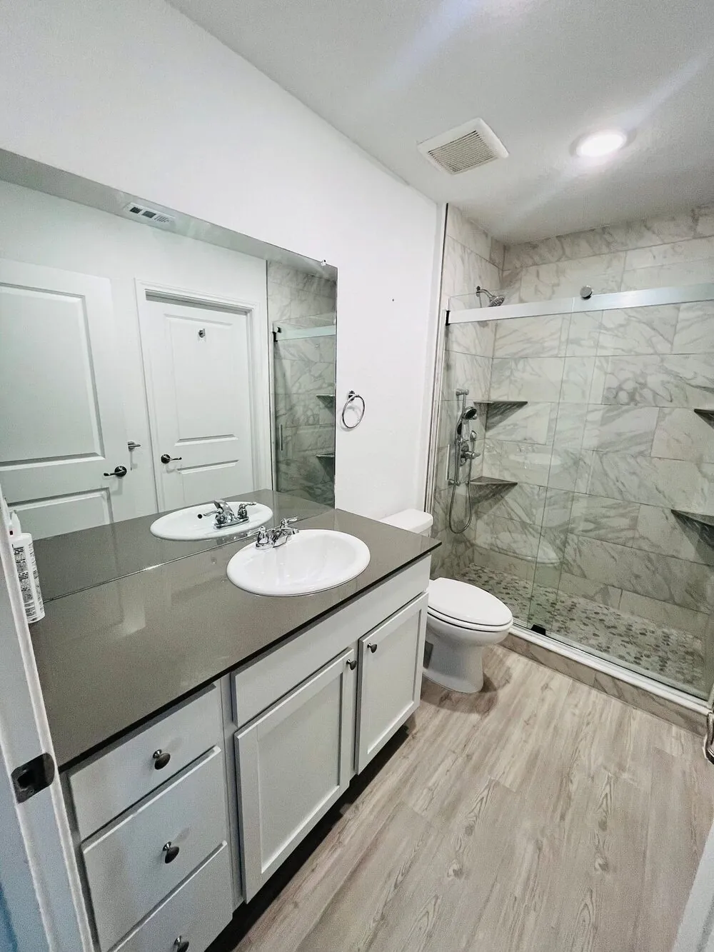 Full bathroom view with a grey-topped vanity, wide mirror and the glass shower beyond