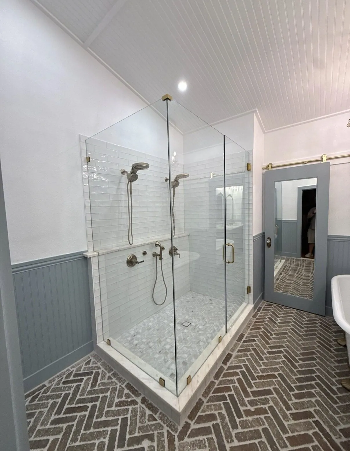 Glass-enclosed shower with white wall tile and a marble mosaic floor, set over herringbone brick-look flooring