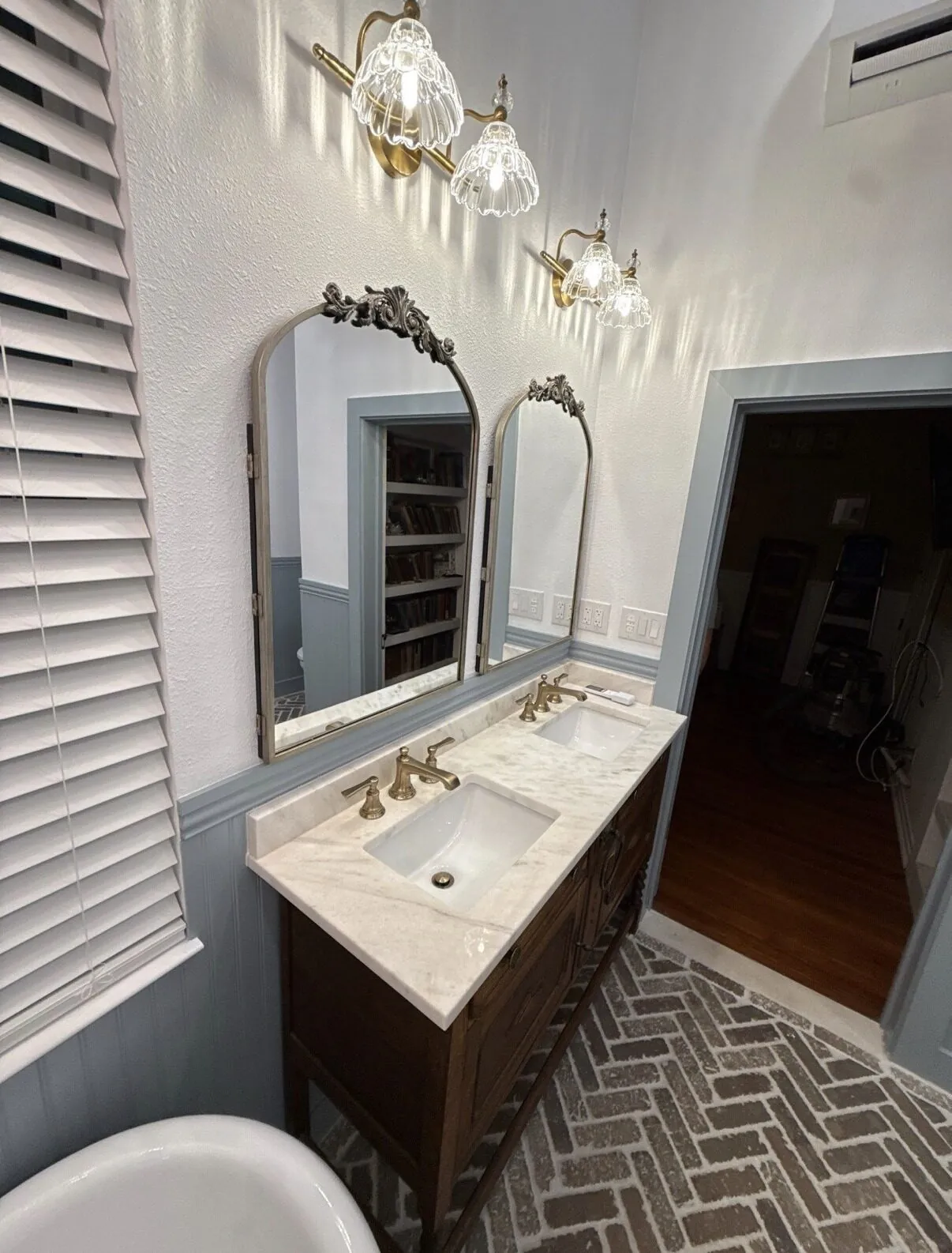 Double vanity with two arched mirrors, marble top and wall sconces beside louvred shutters