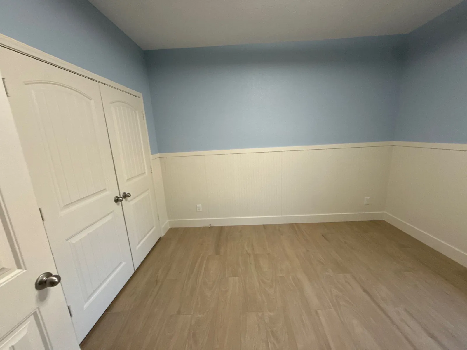 Empty corner of the finished room with white beadboard wainscot below soft blue walls and a wood-look plank floor