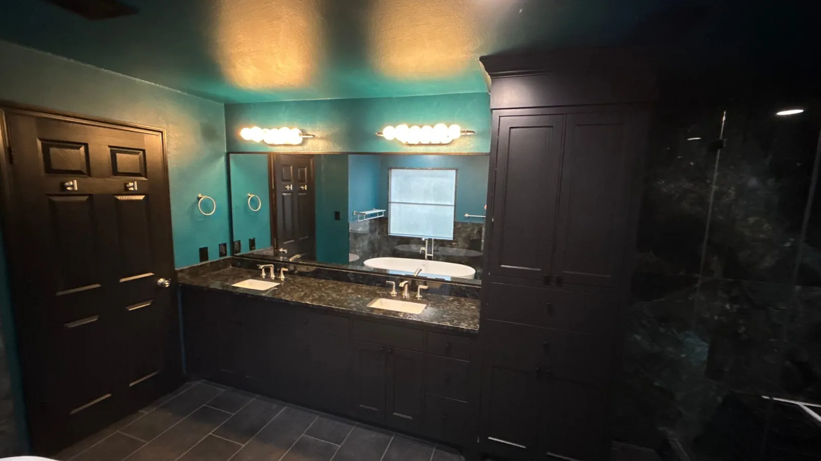 Double vanity with dark cabinetry and stone countertop beneath a full-width mirror and sconce lighting