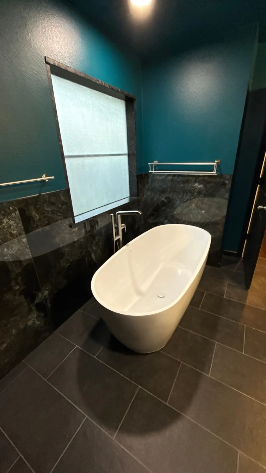 Freestanding soaking tub on dark tile flooring beside its floor-mounted filler